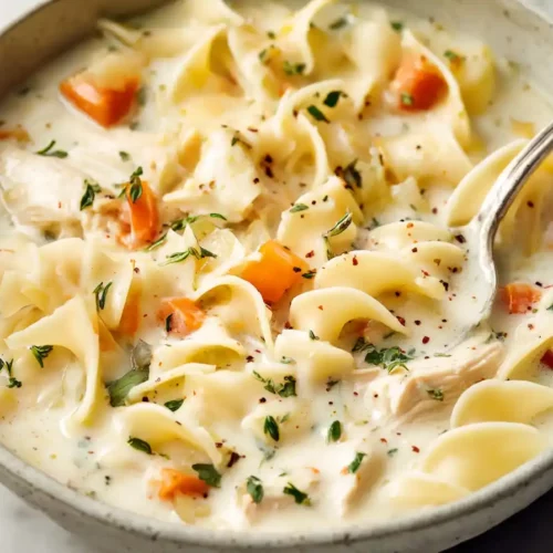 Close-up of a creamy chicken noodle soup served in a rustic bowl with carrots, herbs, and a spoon
